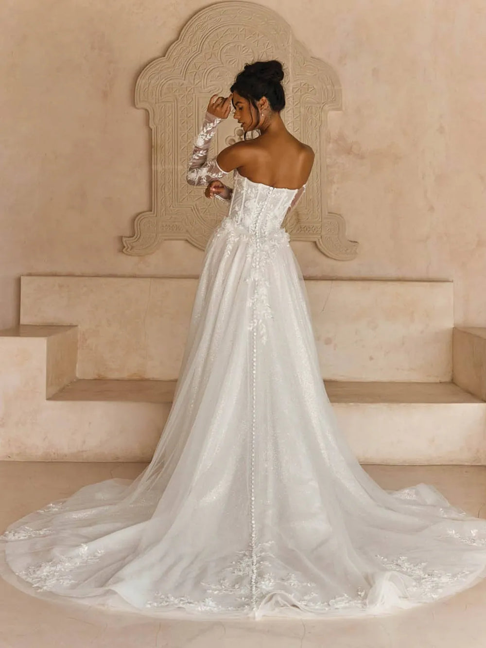 A woman wearing a strapless white wedding gown with long lace sleeves and a dramatic train, photographed in a warm beige architectural setting.