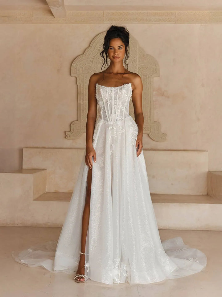 A woman modeling a strapless white wedding gown with a fitted lace bodice, full skirt, and high front slit, standing in a beige architectural setting with a stone throne behind her.