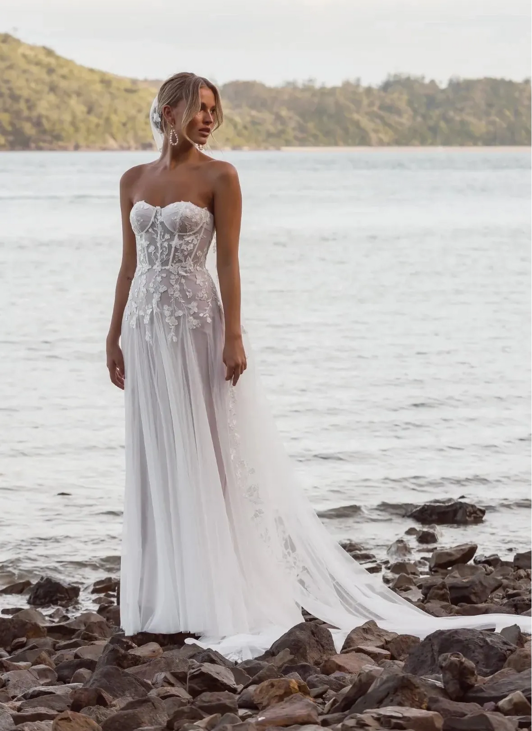 Woman in a strapless wedding dress standing on a rocky shore with water and mountains in the background