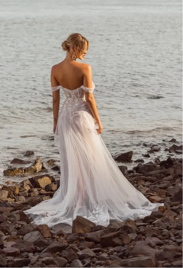 Woman in a wedding dress standing on a rocky beach with water and mountains in the background