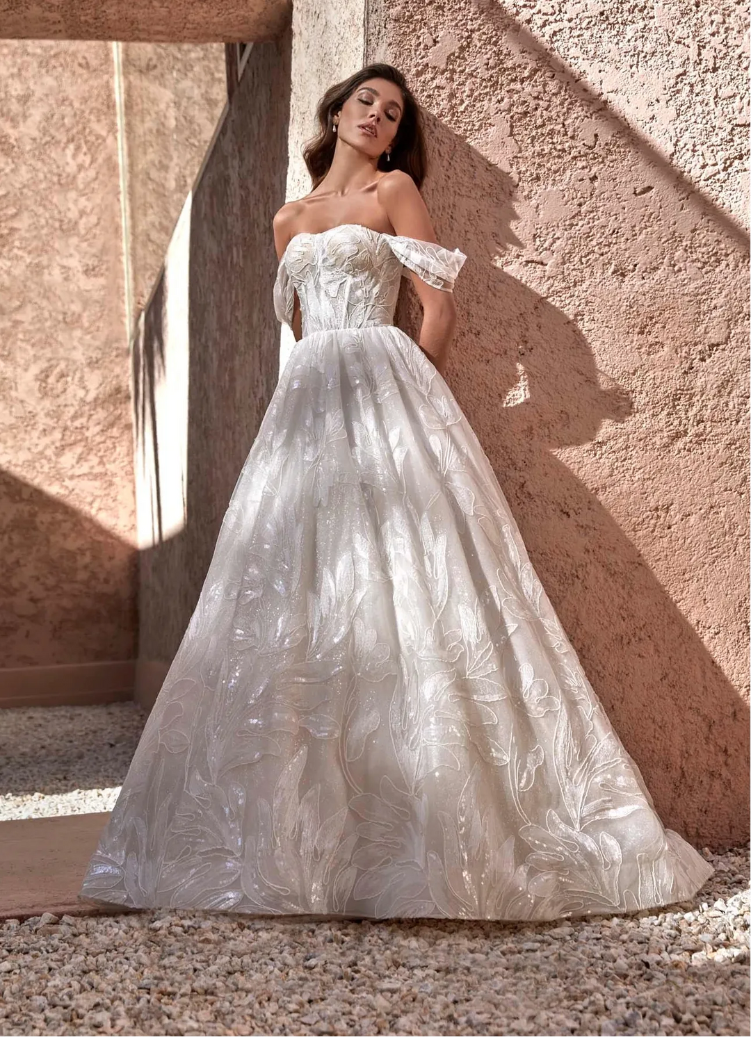 A woman wearing a strapless off-the-shoulder ball gown with a textured, shimmering white and silver tulle skirt, posing against a warm, sunlit stucco wall.