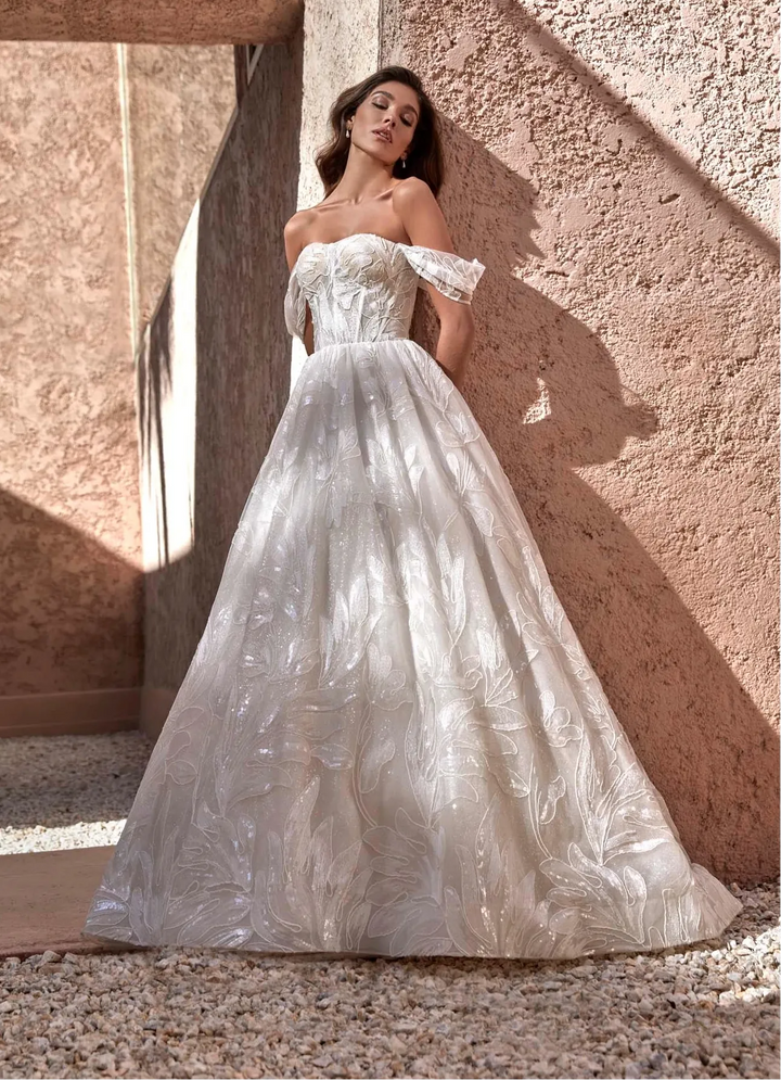 A woman wearing a strapless off-the-shoulder ball gown with a textured, shimmering white and silver tulle skirt, posing against a warm, sunlit stucco wall.