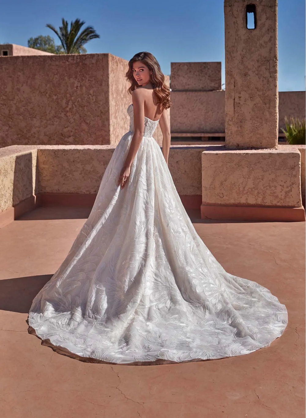 A woman wearing a strapless white lace ball gown with a long train, photographed outdoors against terracotta stucco buildings and a blue sky.