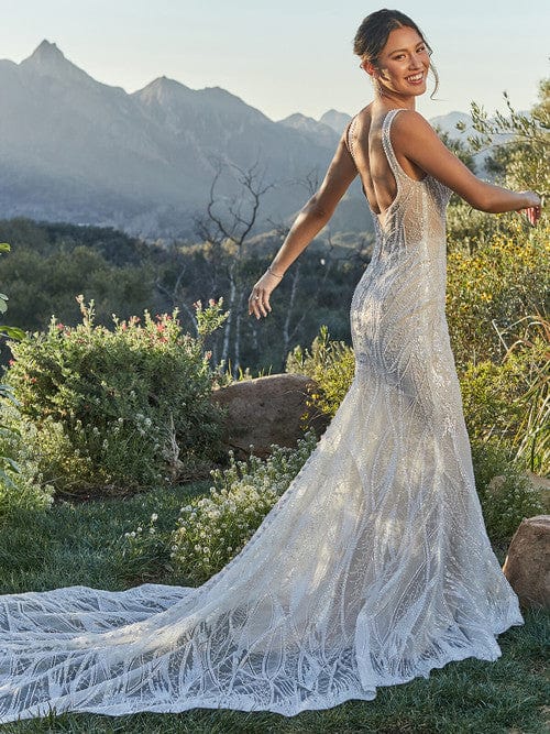 A woman wearing a sleeveless, backless lace wedding gown with a long train stands outdoors in a grassy landscape with mountains in the background.