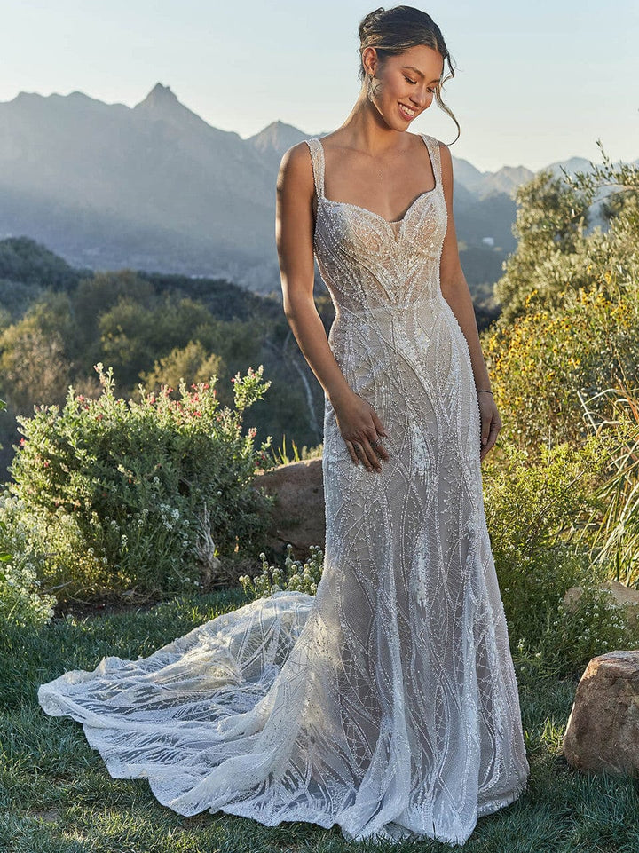 A woman wearing a sleeveless lace wedding gown stands outdoors on grass with mountains and shrubs in the background. The dress has thin straps, a fitted bodice, and a long train.