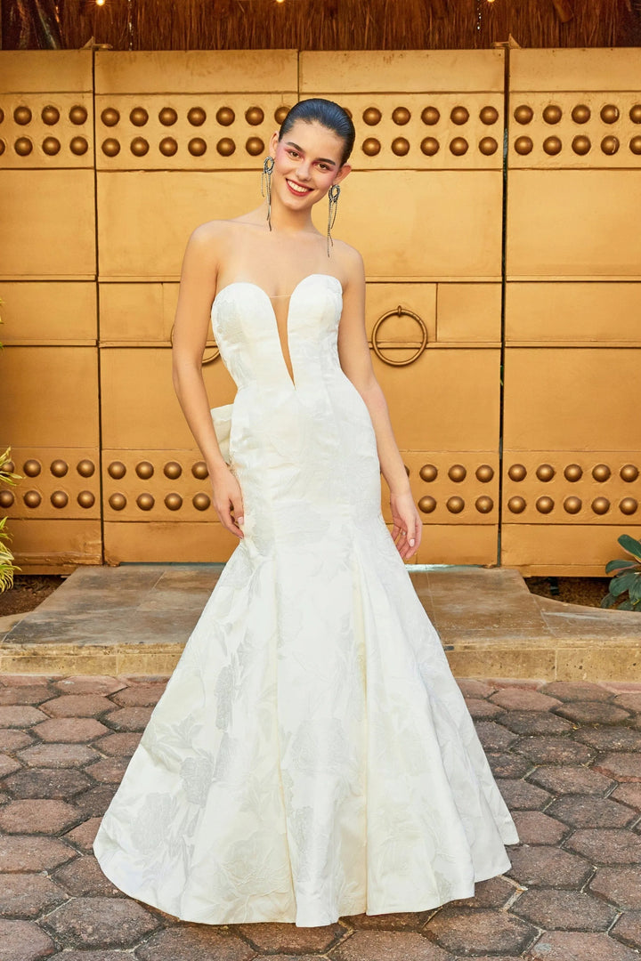 A woman modeling a strapless white mermaid-style wedding gown with a sweetheart neckline, fitted bodice, and full flared skirt, standing in front of a large wooden door.