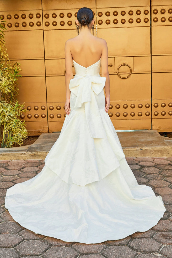 A woman viewed from behind wearing a strapless white wedding gown with a large bow at the back of the waist and a long layered train, standing outdoors in front of a large bronze-toned door.