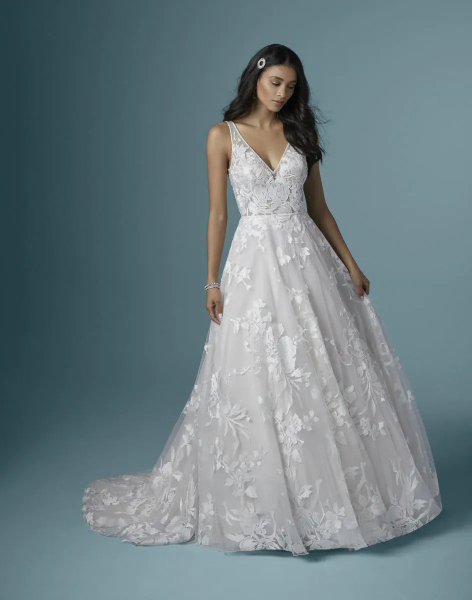 A woman modeling a white sleeveless wedding gown with a deep V-neckline, fitted bodice, and full floral lace skirt against a muted blue-gray studio background.