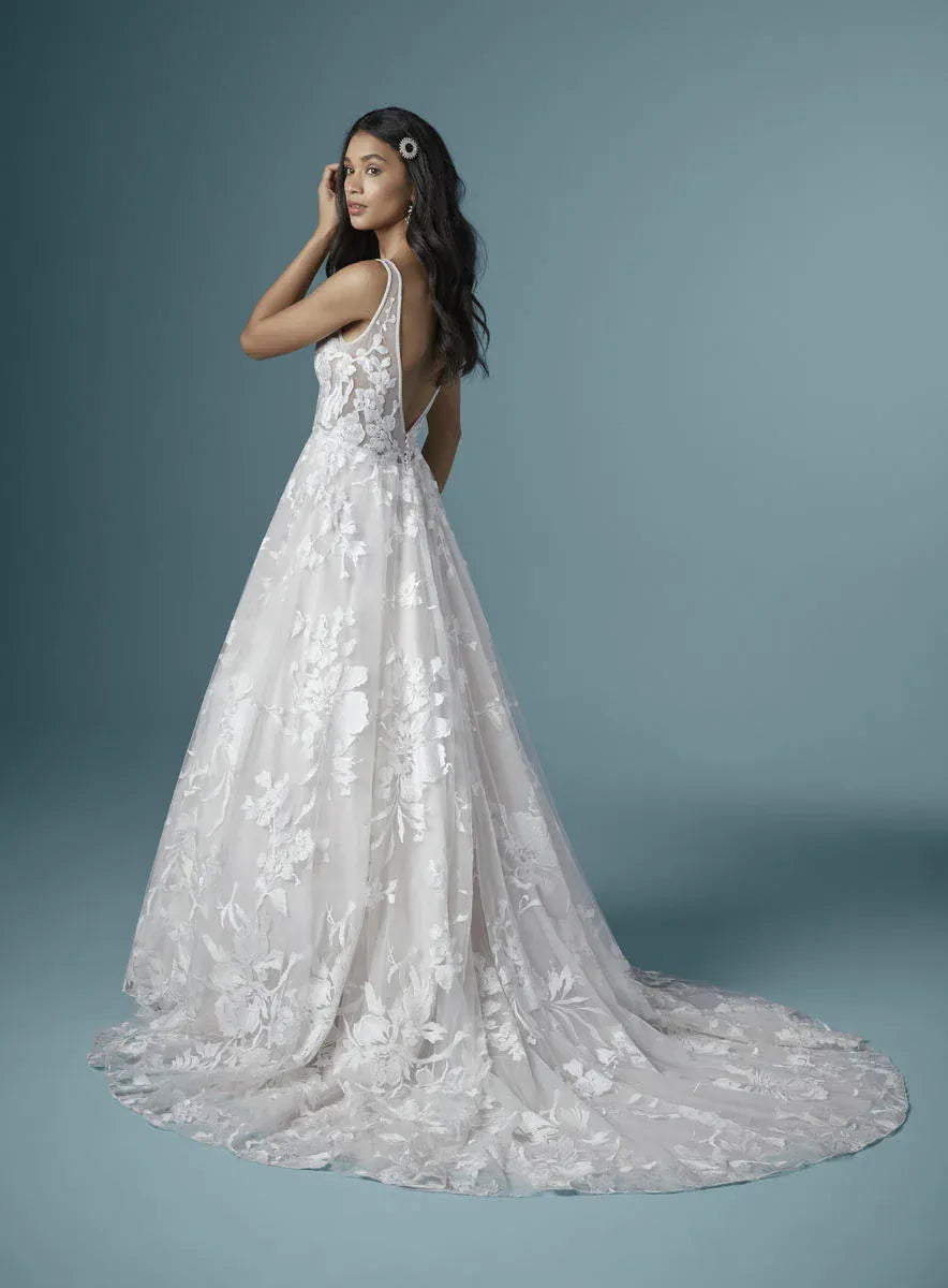 A woman modeling a white lace wedding gown with a fitted bodice, thin straps, and a full flared skirt with a long train, photographed against a soft blue-gray studio background.