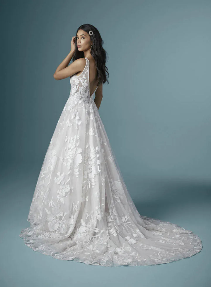 A woman modeling a white lace wedding gown with a fitted bodice, thin straps, and a full flared skirt with a long train, photographed against a soft blue-gray studio background.