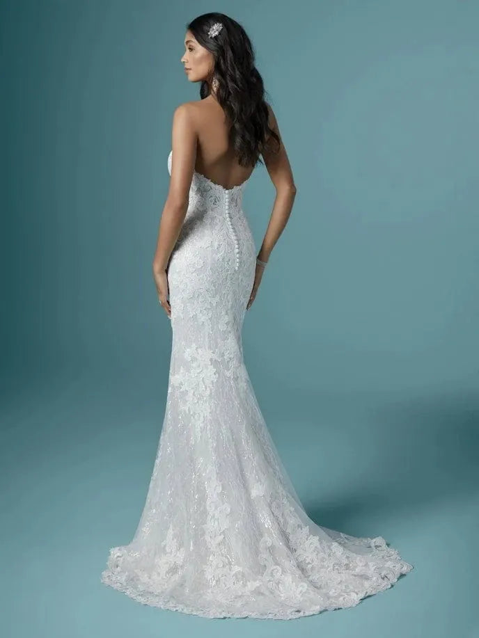 A woman modeling a white lace mermaid-style wedding dress with a low open back and long train, photographed against a smooth blue studio backdrop.