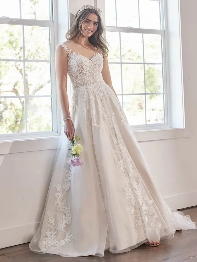 A woman wearing a sleeveless lace wedding gown with a fitted bodice and full tulle skirt, standing indoors near large white-framed windows and holding a small bouquet.