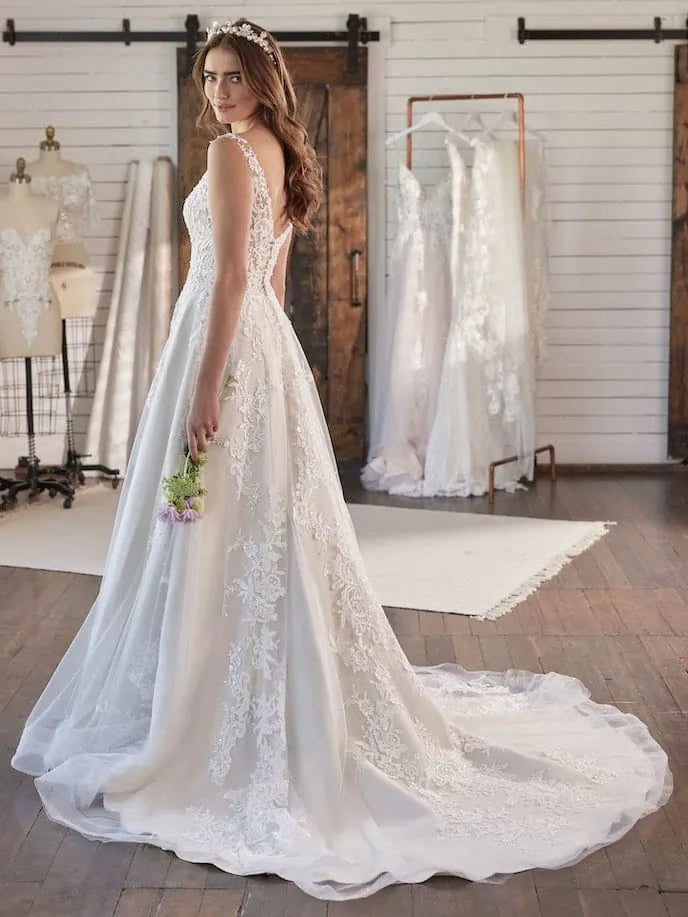 A woman wearing a white lace and tulle wedding gown stands in a bridal shop, holding a small bouquet. The dress has a fitted bodice, sheer lace panels, and a long train.
