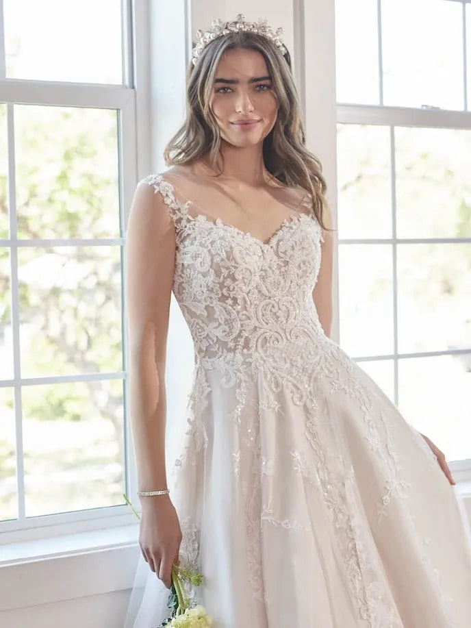 A woman wearing a sleeveless lace wedding gown with a fitted bodice and full skirt, standing indoors near large bright windows and holding a small bouquet.