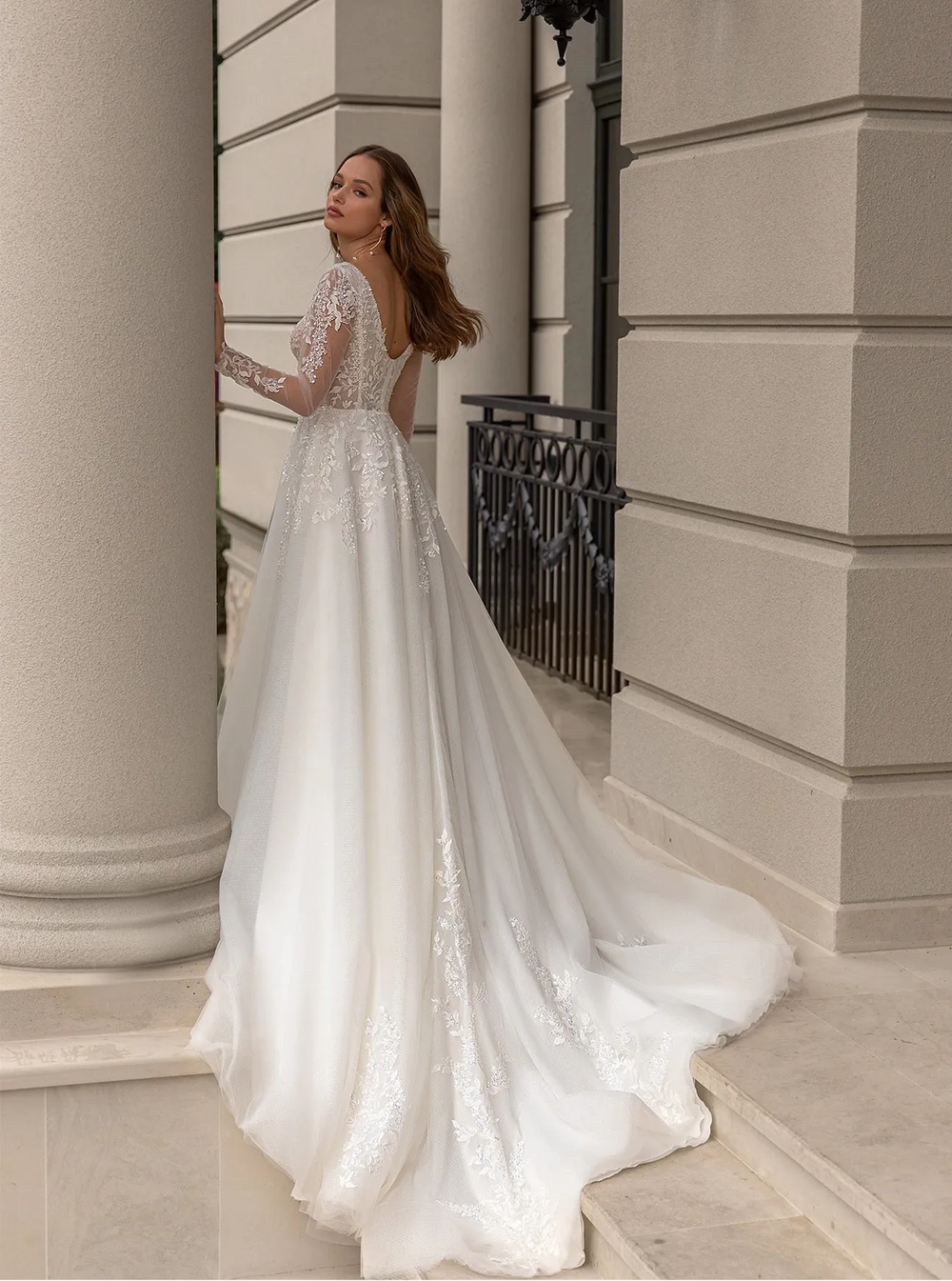 A woman wearing a white lace wedding gown with a long train, standing outdoors in front of a beige stone building with columns and a black wall lamp.