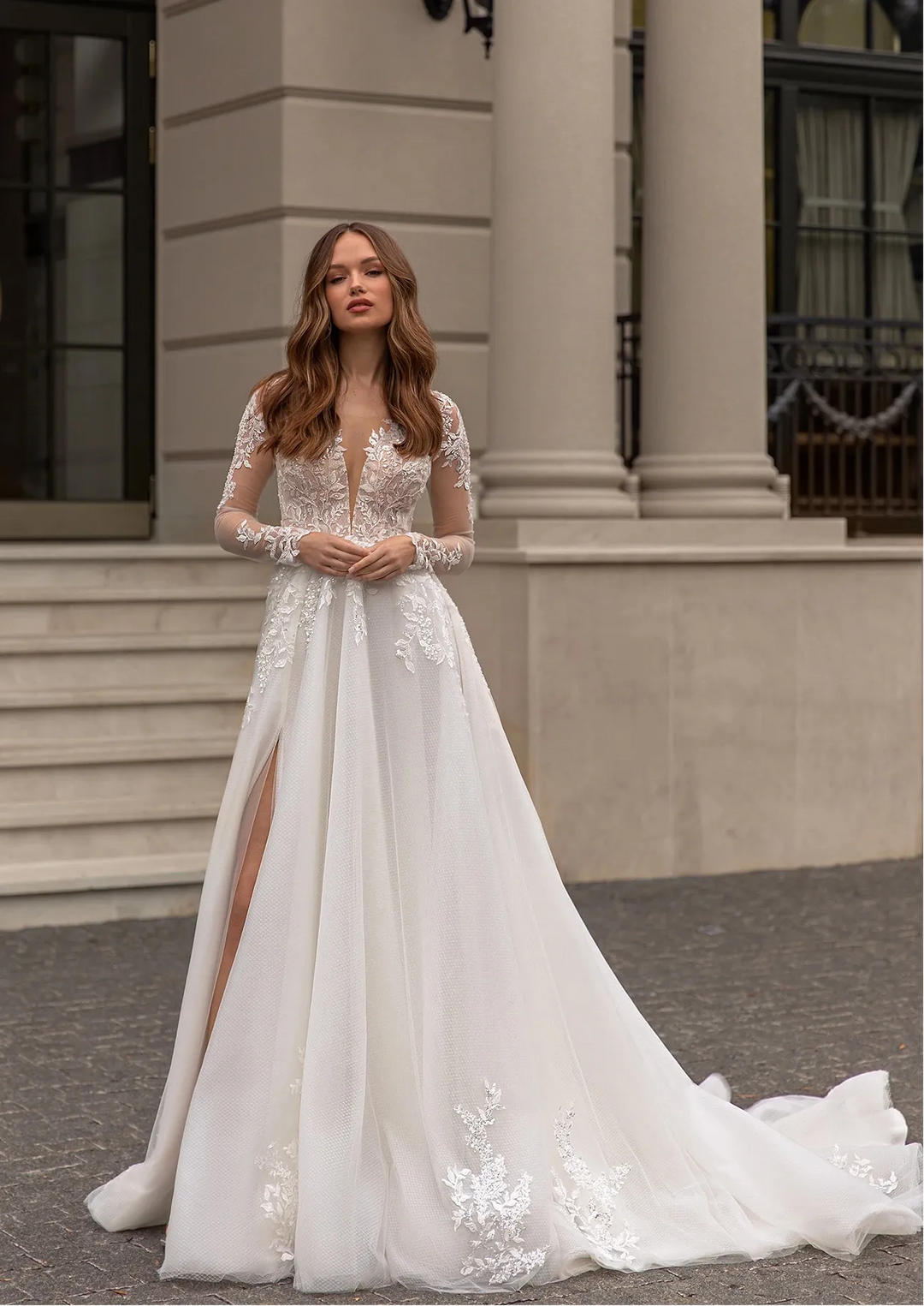 A woman wearing a long-sleeved white wedding gown with lace and tulle details, standing outdoors in front of a classical stone building with columns.