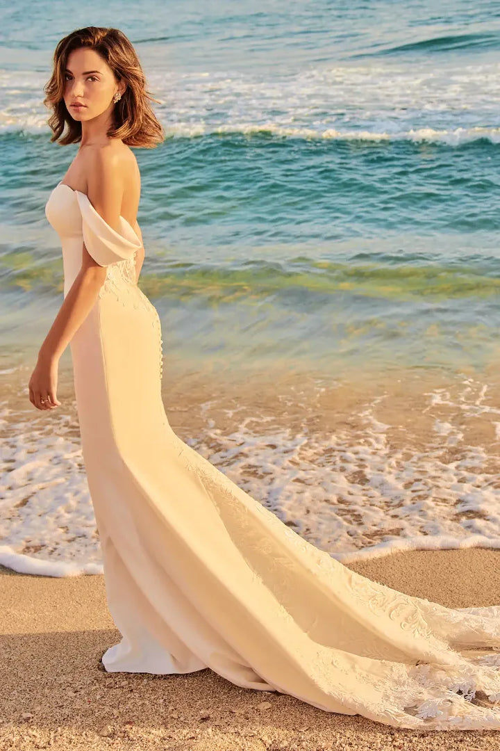 A woman wearing a strapless off-the-shoulder floor-length gown with a long train stands on a sandy beach with ocean waves in the background.