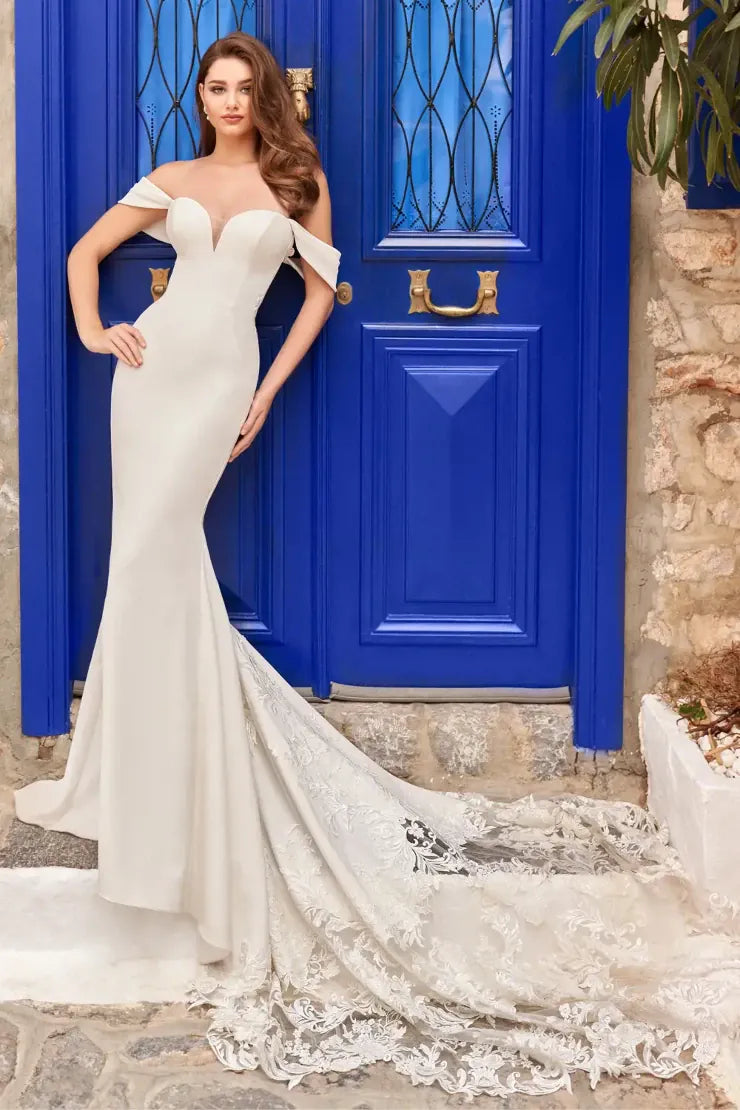 An off-the-shoulder ivory wedding gown with a sweetheart neckline and a dramatic lace-trimmed train, worn by a model standing in front of a vibrant blue door.