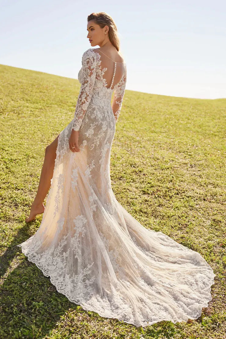 A woman wearing a long-sleeve lace wedding gown stands outdoors on a grassy hill, photographed from the back as the dress train spreads across the lawn under a bright sky.