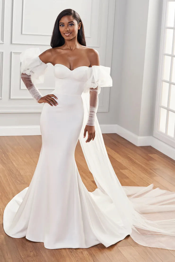 A woman modeling an off-the-shoulder white mermaid-style wedding gown with sheer long sleeves, a sweetheart neckline, and a long flowing train in a bright indoor room with wood floors.