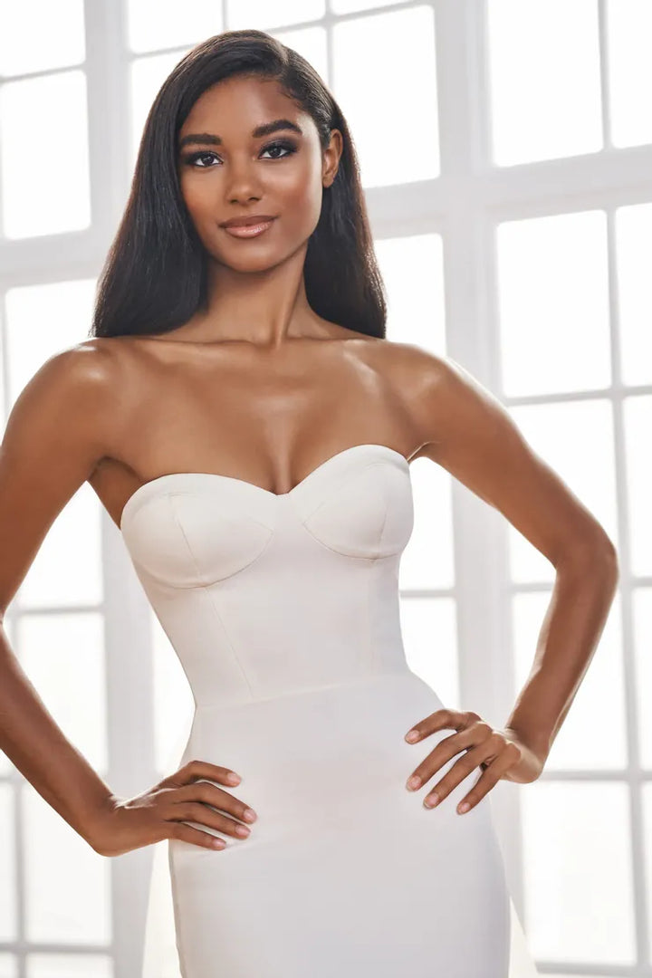 A woman modeling a strapless sweetheart-neckline white wedding dress with structured underbust padding, standing in front of a bright white window grid.