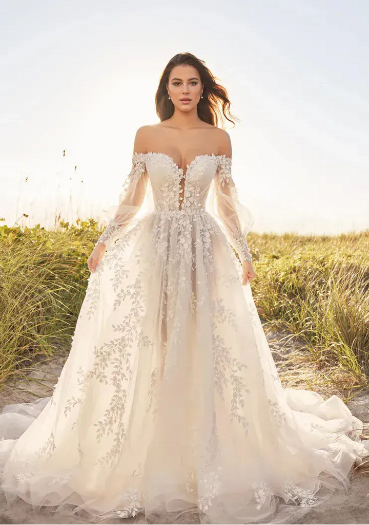 Woman in a white lace wedding dress standing in a field with a clear sky.
