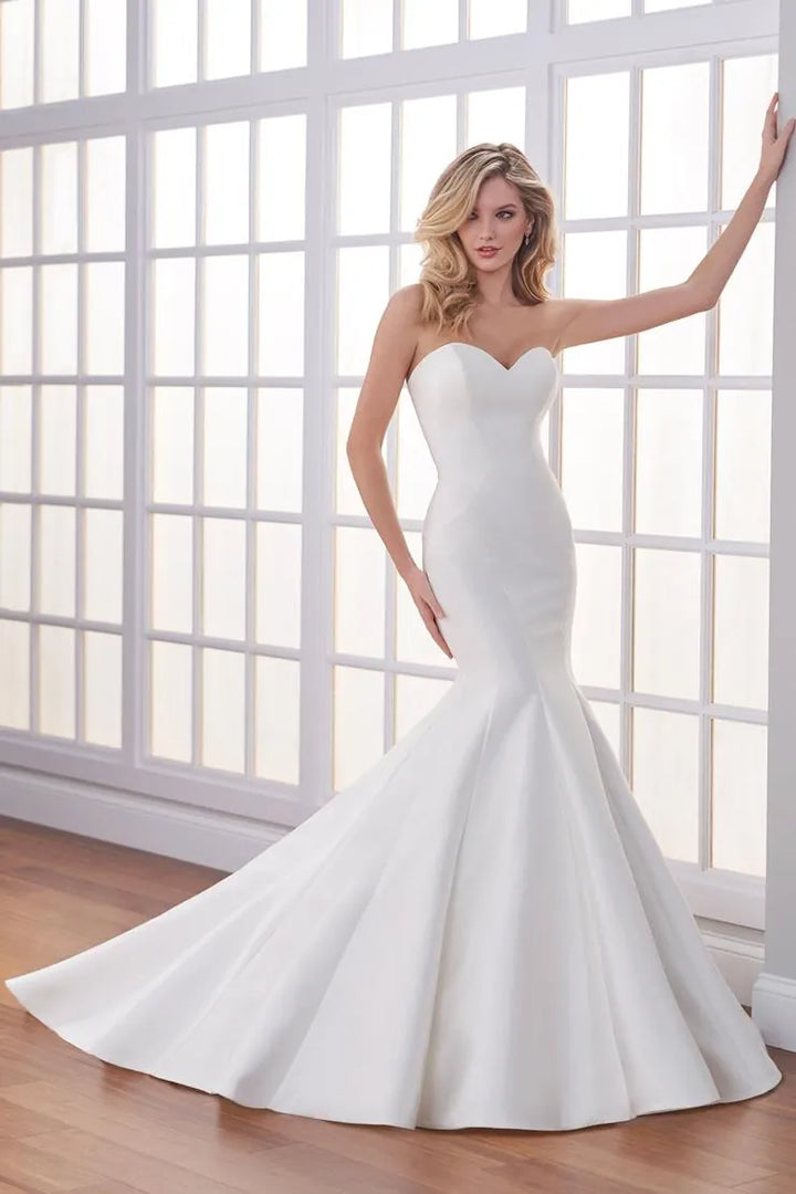 A woman models a strapless white mermaid-style wedding gown with a sweetheart neckline, fitted bodice, and flared skirt, posed in front of large white-framed windows on a wood floor.