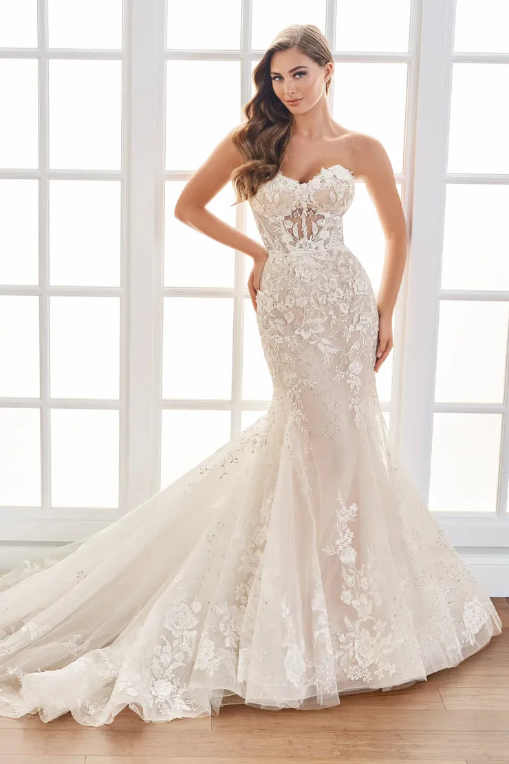 A woman modeling a strapless mermaid-style wedding gown with a sweetheart neckline, fitted lace bodice, and full tulle skirt with floral lace appliqué, standing in a bright room with large white window panes and a wood floor.