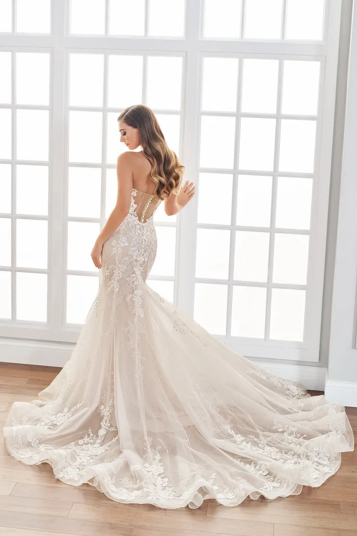 A woman wearing a strapless ivory wedding gown with lace applique and a long train, standing in front of a bright white window.