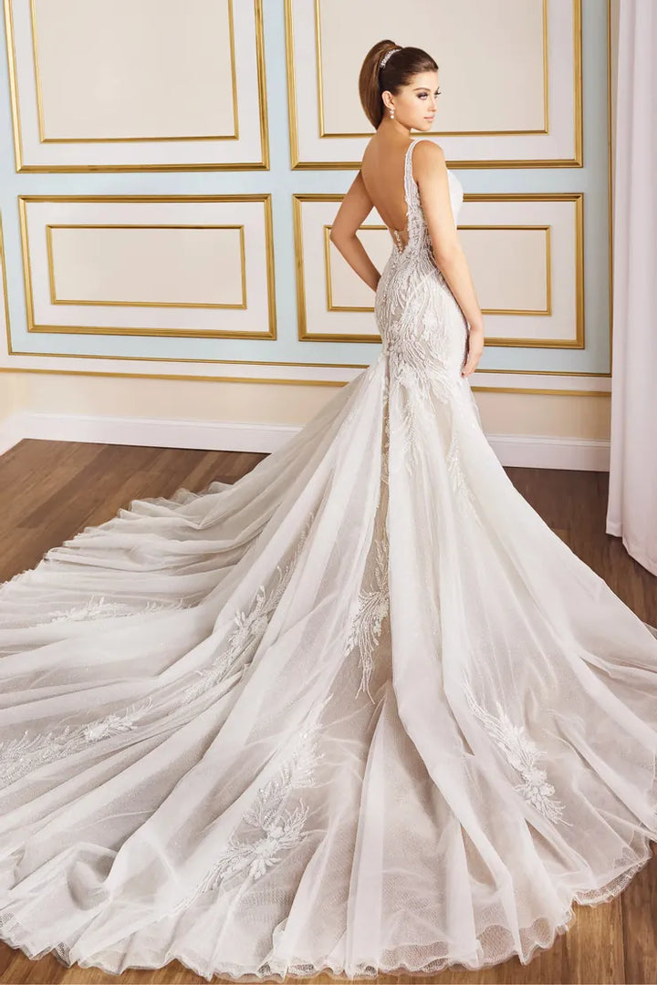 A woman modeling a bridal gown from the back, showing a fitted lace mermaid silhouette, deep open back, and a very full sheer tulle train on a wood floor in an elegant room.