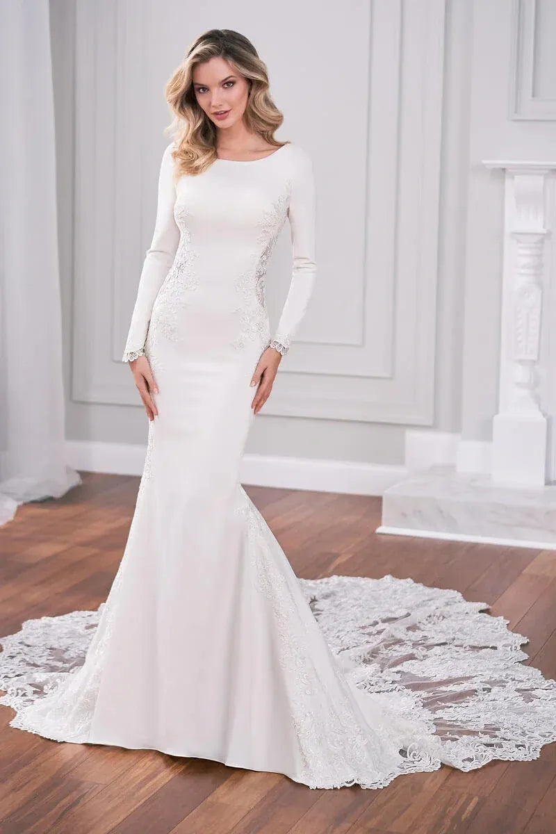 A woman modeling a long-sleeve white mermaid-style wedding gown with lace detailing and a train in an elegant indoor room with wood floors and white paneled walls.