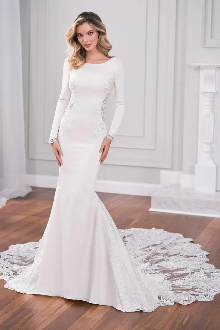 A woman modeling a long-sleeve white mermaid-style wedding gown with lace detailing and a train in an elegant indoor room with wood floors and white paneled walls.