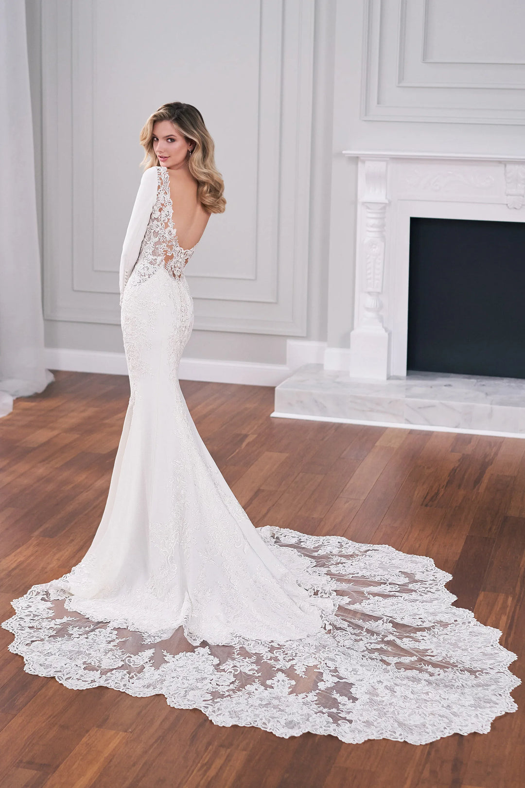 A woman modeling a white mermaid-style wedding dress with a long lace train in an elegant room with wood floors and a white fireplace.