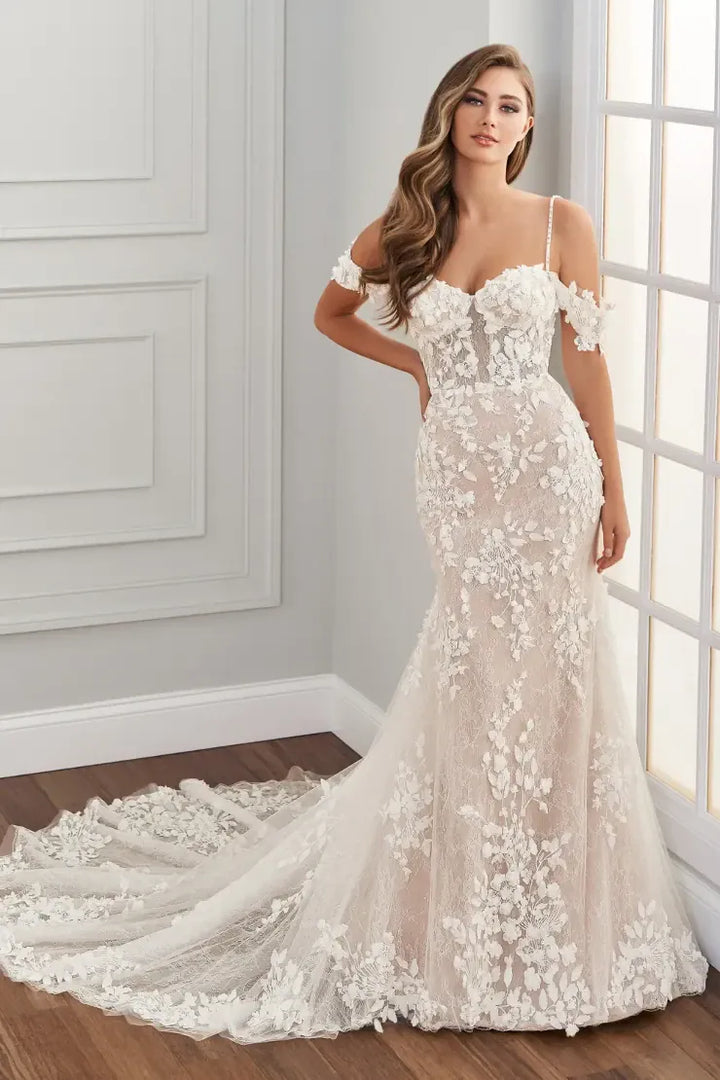 A woman modeling an off-the-shoulder mermaid-style wedding gown with floral lace applique and a long train in a bright indoor room with paneled walls and a window.