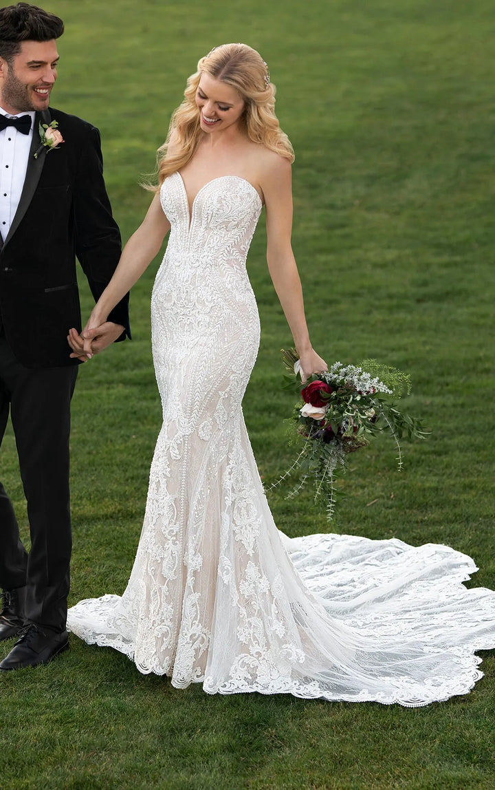 Woman in a white lace wedding dress holding hands with a man in a black suit on a grassy field.