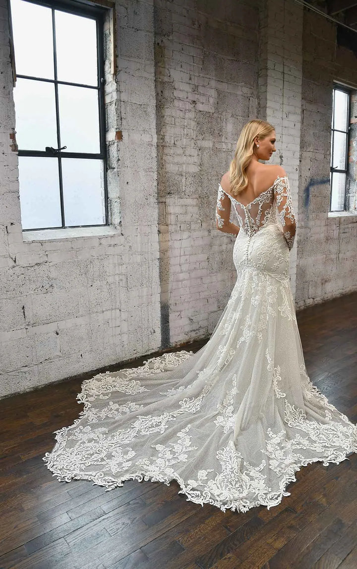 A woman wearing a white lace mermaid-style wedding gown stands in a loft-style room with exposed brick walls and large windows, showing the back of the dress with its long train.