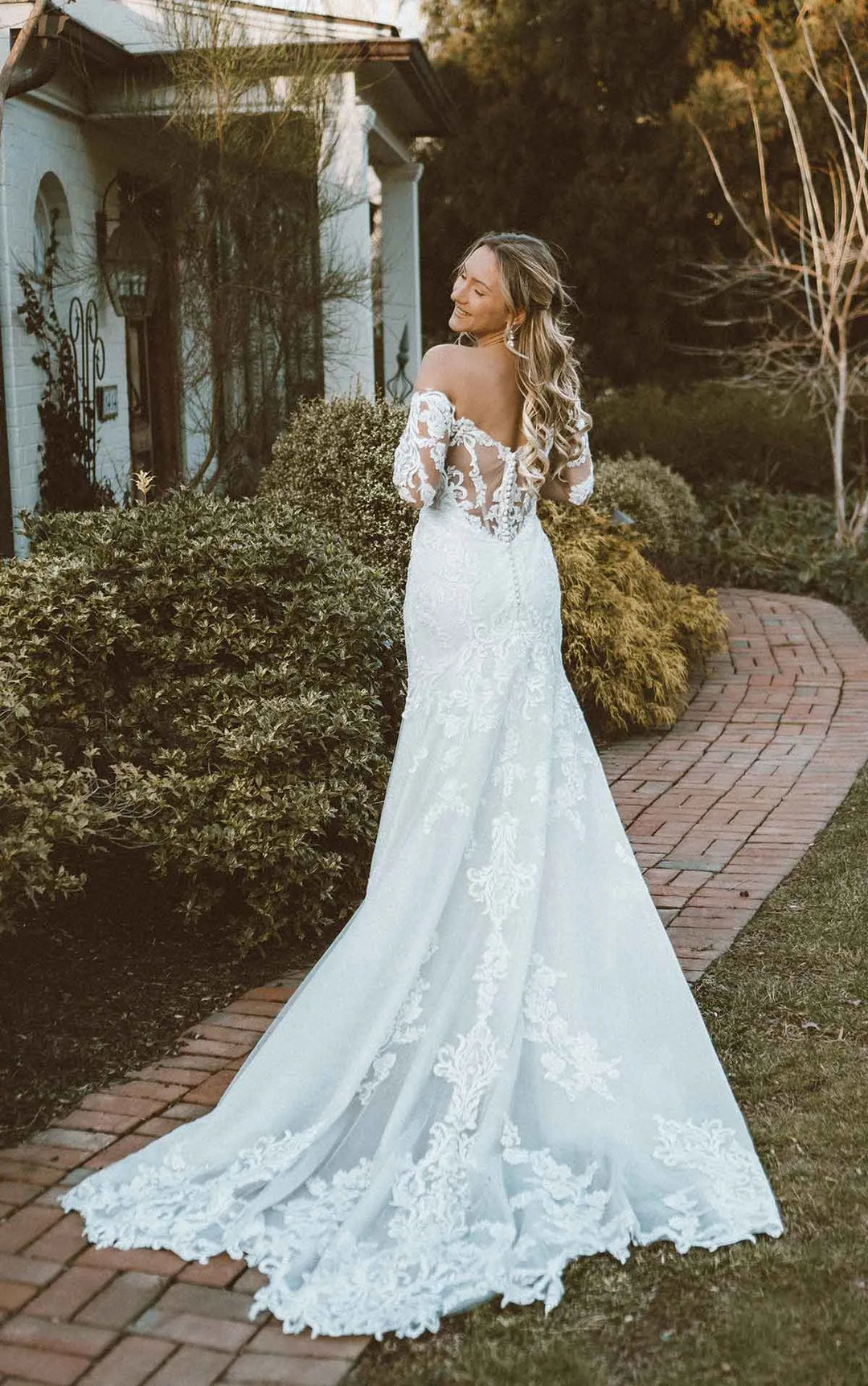 A woman wearing a white off-the-shoulder lace wedding gown stands on a brick path outdoors, with shrubs, trees, and a house visible in the background.