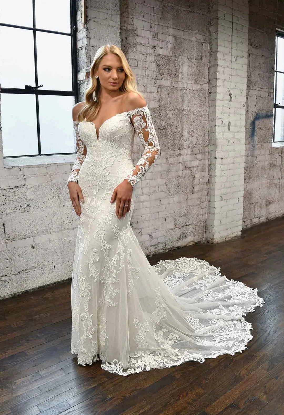 A woman modeling an off-the-shoulder lace wedding gown with long sheer lace sleeves and a full train, standing in a rustic brick warehouse interior with wood floors.