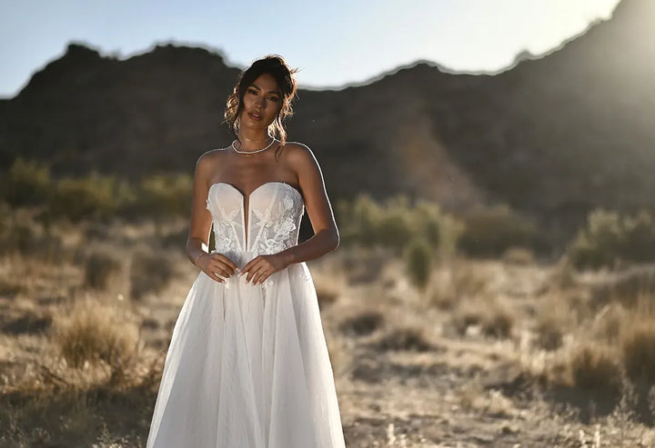 A woman wearing a strapless white wedding gown stands outdoors in a desert landscape with mountains in the background. The dress has a fitted lace corset-style bodice and a flowing tulle skirt.