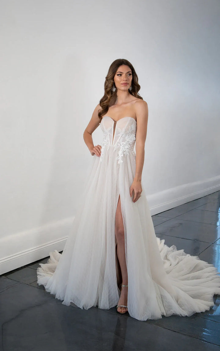 A woman models a strapless white wedding gown with a sweetheart neckline, lace applique on the bodice, a thigh-high slit, and a voluminous tulle train on a dark tiled floor against a white wall.