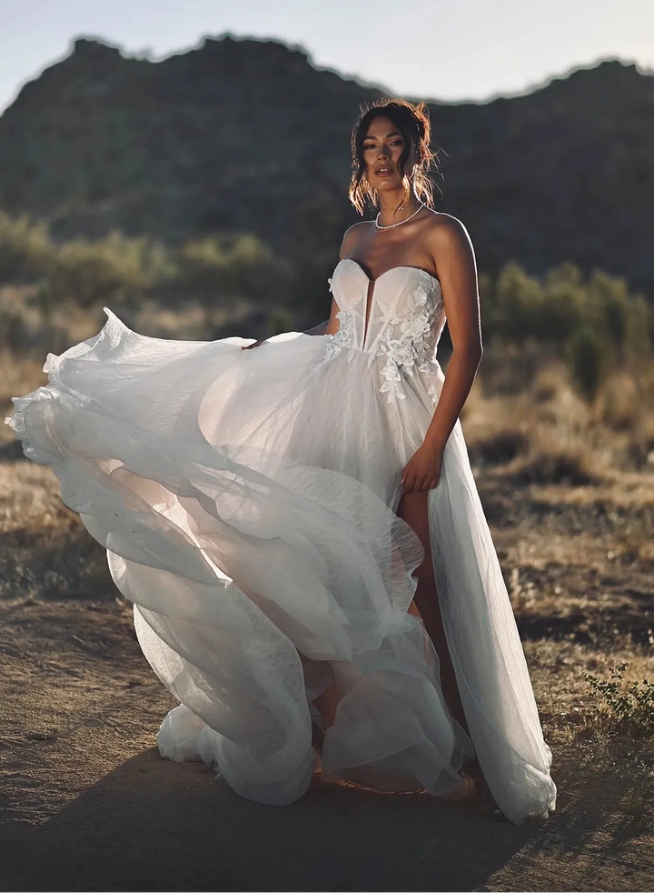 A woman wearing a strapless white wedding gown with a voluminous flowing skirt stands outdoors in a dry, mountainous landscape. The dress has a fitted bodice with lace or embroidered detailing and a high slit, with the skirt being blown slightly by the wind.
