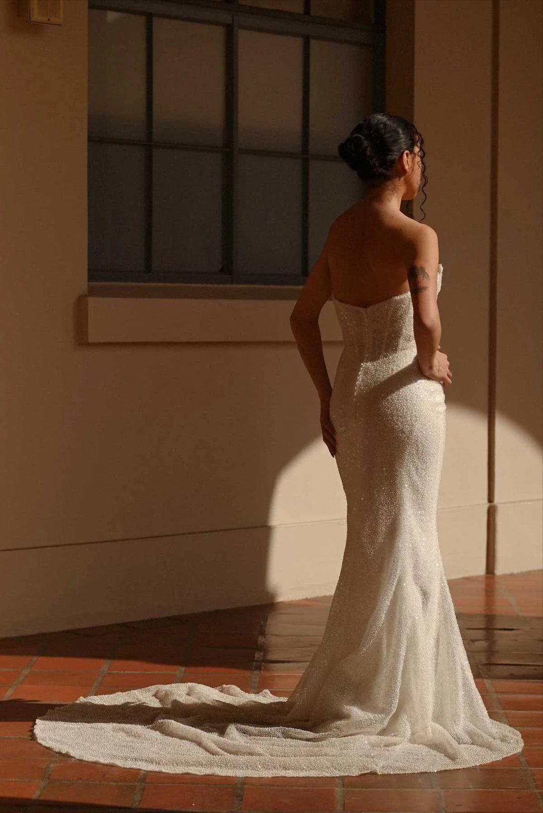 Woman in a white strapless gown standing in a sunlit room with a window in the background.