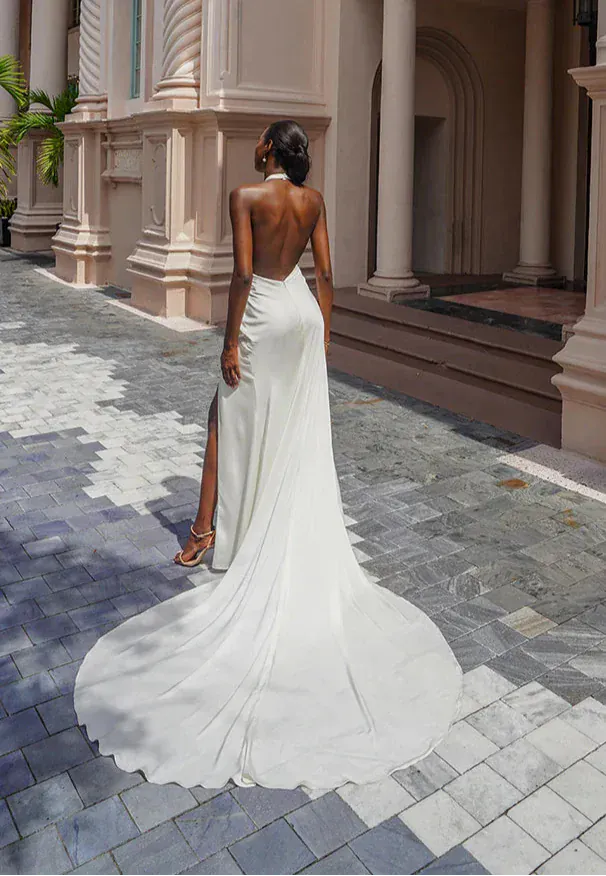 A woman stands outdoors in front of a building with large columns, wearing a white backless wedding gown with a long train and a high slit on one leg.
