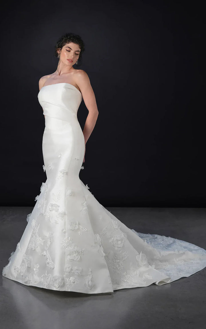 Woman wearing a strapless white wedding dress with floral details on a dark background