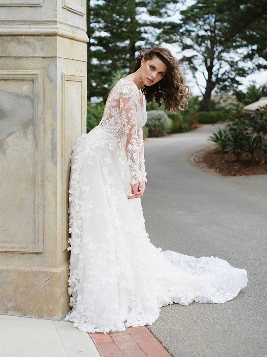 A woman wearing a white lace wedding gown with a long train, leaning against a stone pillar in an outdoor garden setting.