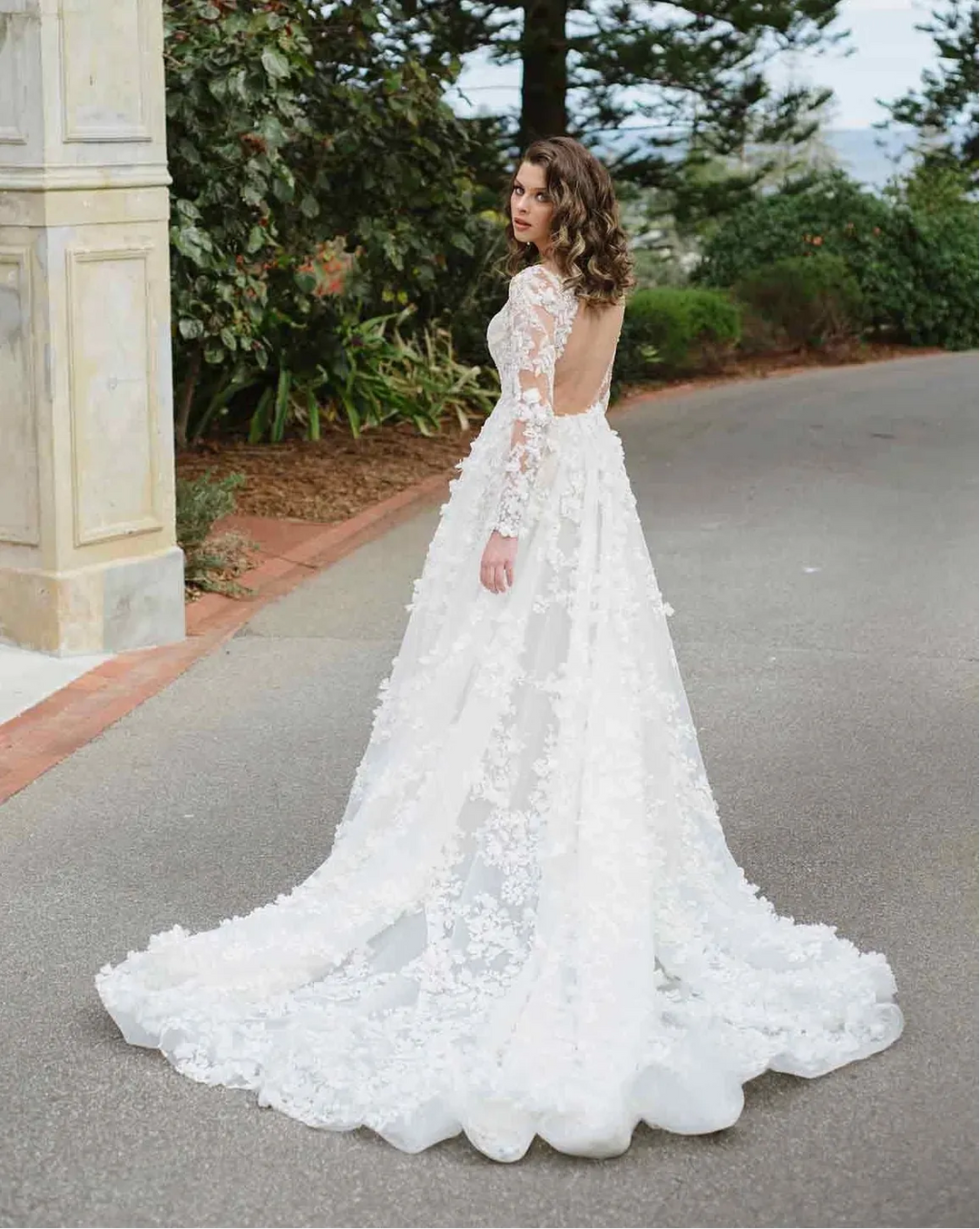 A woman wearing a white lace wedding gown stands outdoors on a paved road with trees and a stone pillar in the background, photographed from behind to show the back of the dress.