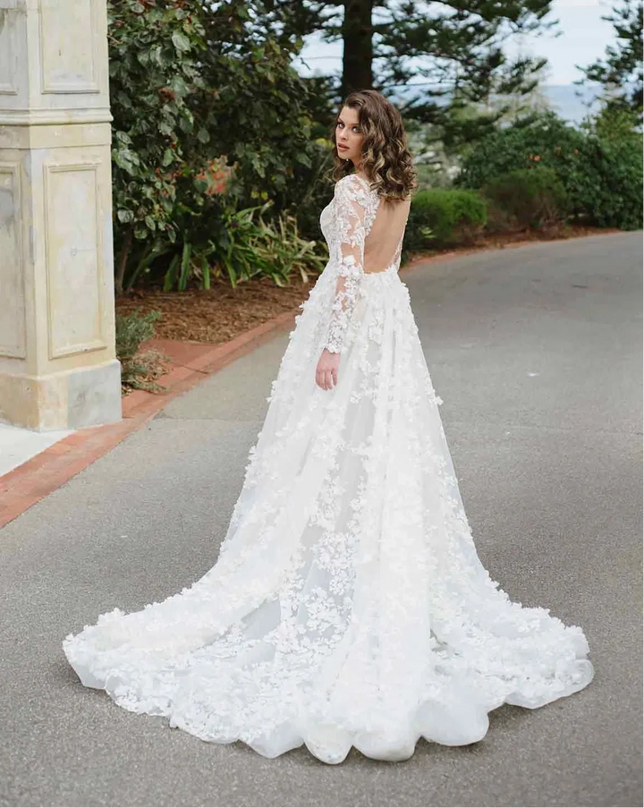 A woman wearing a white lace wedding gown stands outdoors on a paved road with trees and a stone pillar in the background, photographed from behind to show the back of the dress.