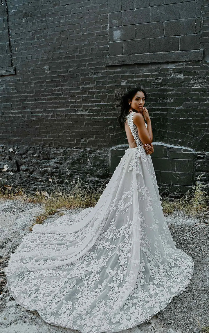 A woman wearing a sleeveless lace wedding gown with a long train stands outdoors against a dark brick wall, looking back over her shoulder.