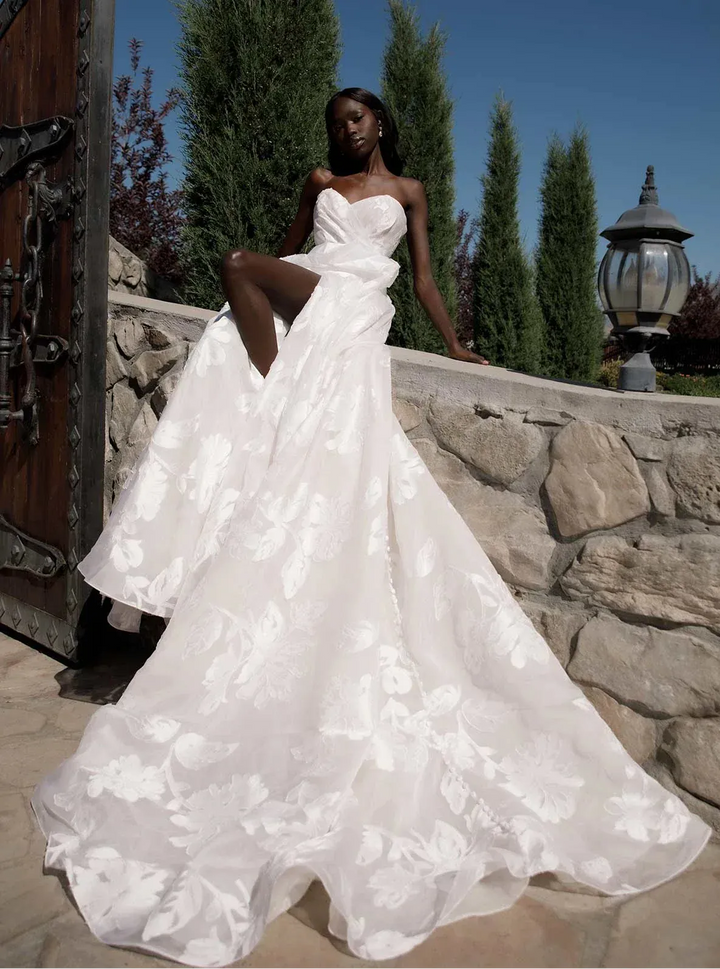 Woman in a white wedding dress standing outdoors with stone wall and trees in the background
