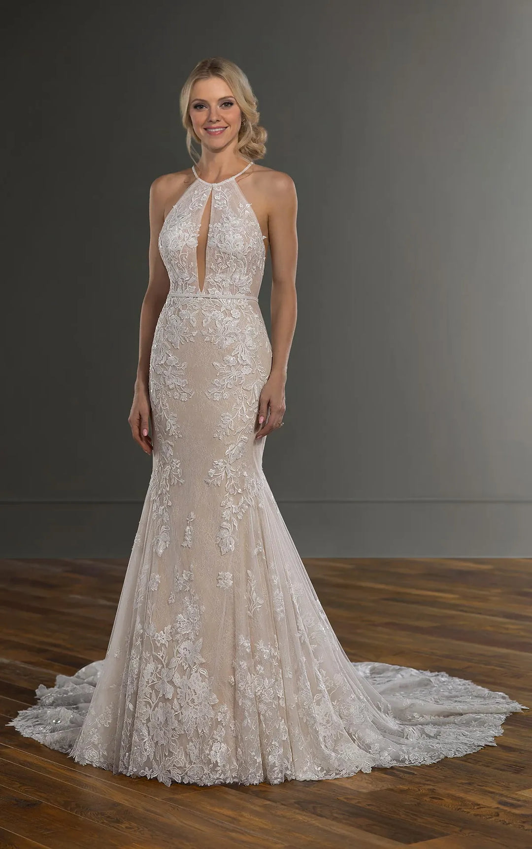 A woman modeling a sleeveless halter-neck wedding gown with a fitted silhouette, floral lace overlay, and a long train on a dark gray studio background with a wood floor.